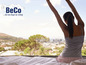 Une femme s'étire au lit avec vue sur une ville, avec le logo BeCo.