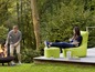 Une femme assise dans un fauteuil Fatboy vert avec un repose-pieds, tandis qu'un homme ajoute du bois à un brasero.
