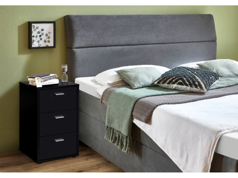 Une table de chevet noire avec trois tiroirs à côté d'un lit gris avec une couette blanche.