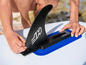 Une main fixant une aileron noir sur un paddleboard gonflable.