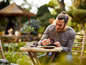 Un homme assis à une table de jardin, examinant un petit modèle de bateau, à côté d'un bonsaï.