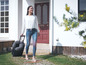 Une femme avec une valise à roulettes et un coussin de nuque devant une maison.