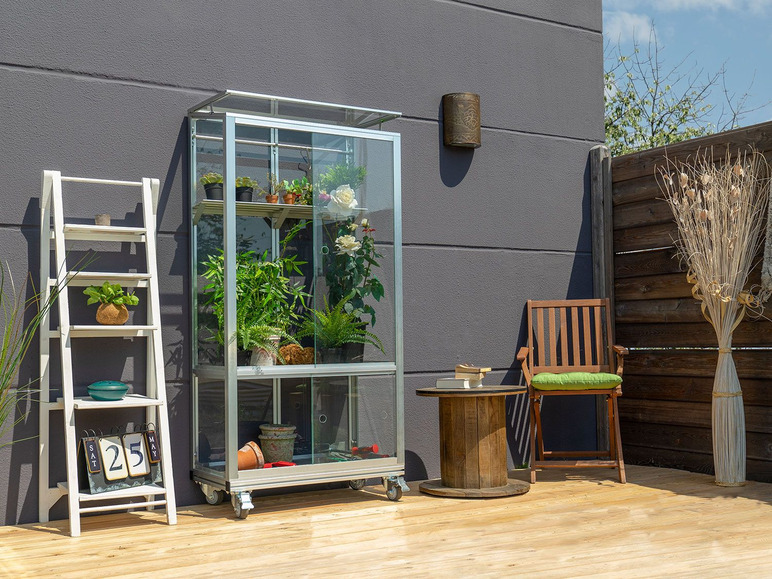 Une serre en verre avec des plantes, une chaise en bois et une table en bois sur une terrasse.