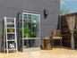 Une serre en verre avec des plantes et une chaise en bois avec un coussin vert sur une terrasse.