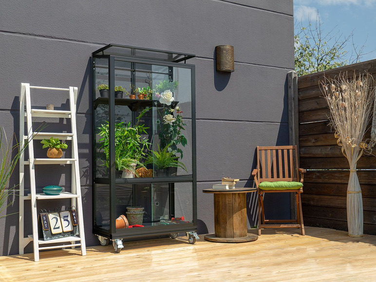 Une serre en métal noir avec différentes plantes sur roulettes, une chaise en bois et une table en bois sur une terrasse en bois.