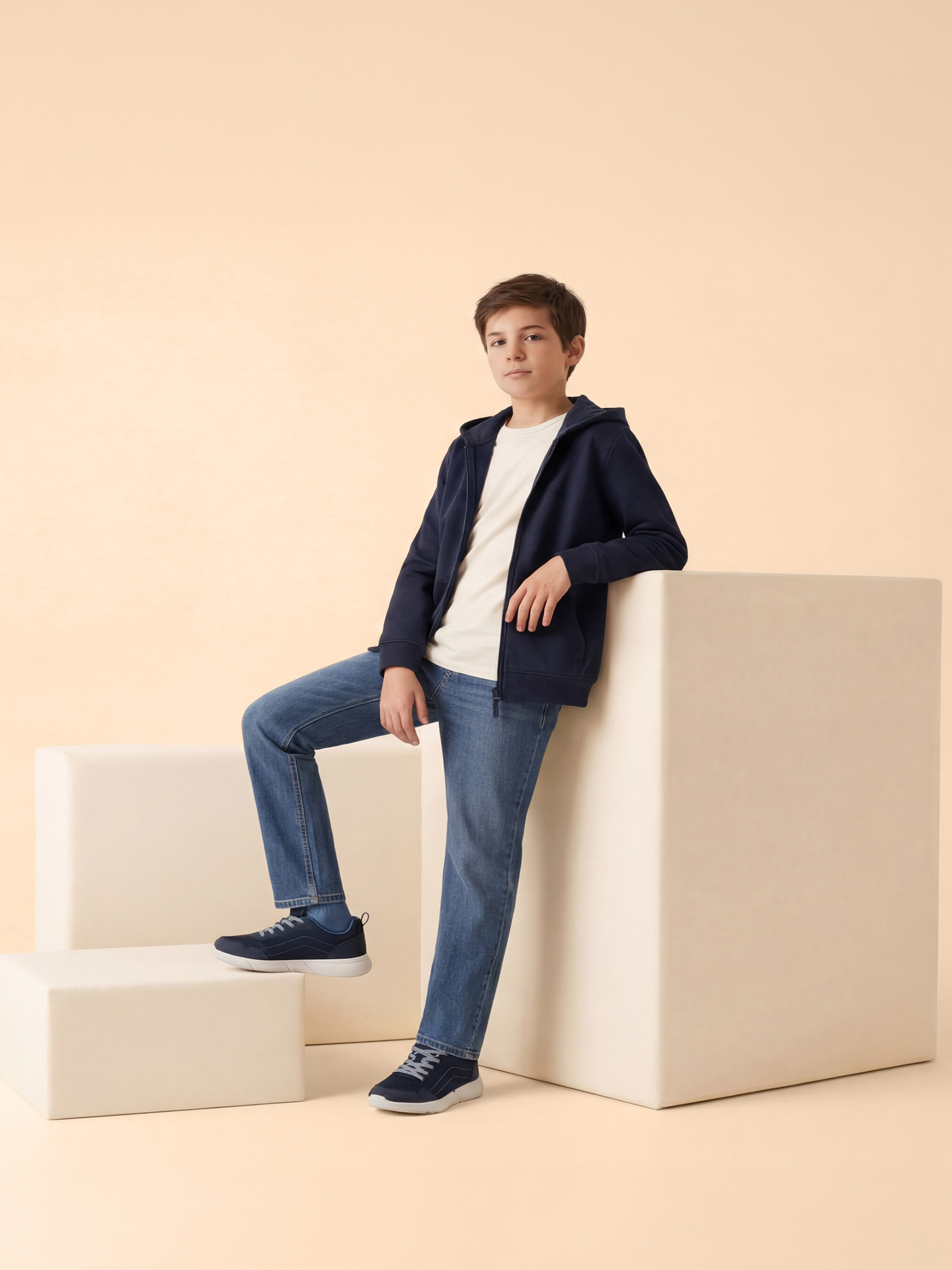 Garçon en sweat à capuche bleu, t-shirt blanc et jean bleu, appuyé sur des blocs blancs.