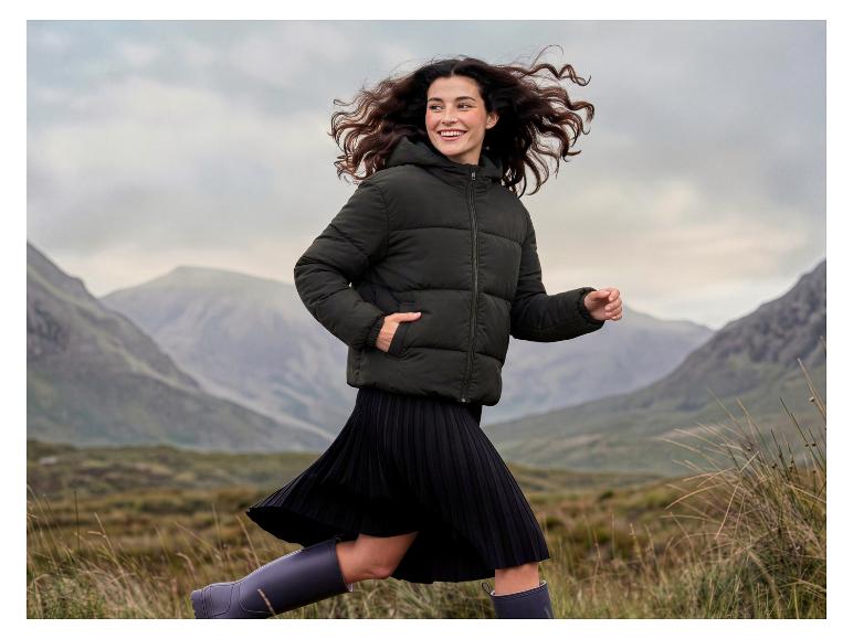 Femme en doudoune vert foncé, jupe plissée noire et bottes en caoutchouc, courant en extérieur.