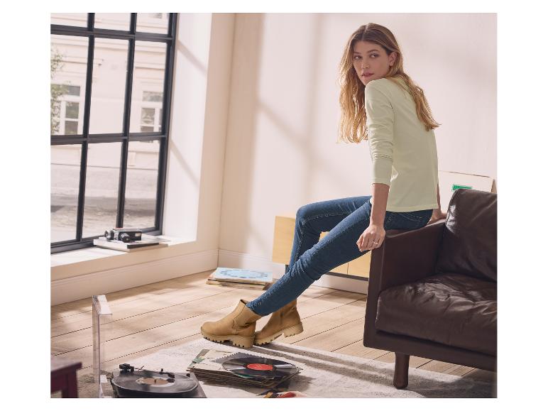 Femme en pull vert clair et jean assise sur un canapé en cuir, avec des bottines beiges.