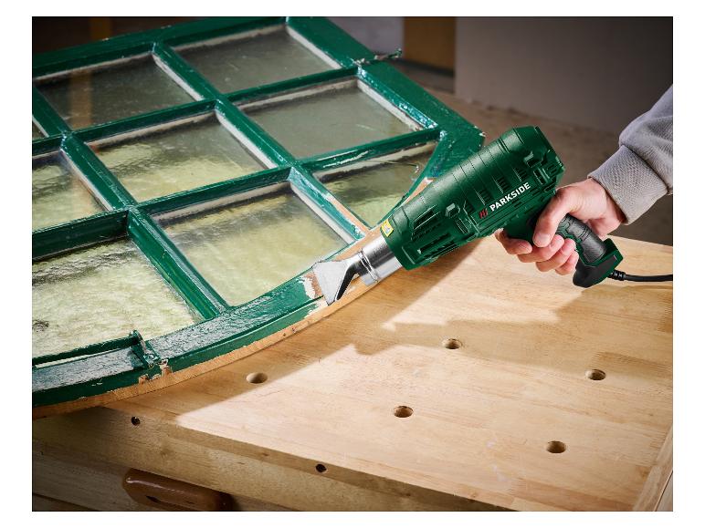 Une personne utilise un décapeur thermique Parkside pour enlever la peinture d'un cadre de fenêtre vert.