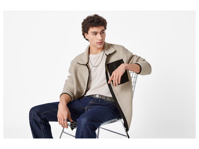Homme assis sur une chaise, portant une veste polaire beige, un T-shirt clair et un jean.