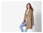 Femme en manteau brun et jeans posant près d'un tabouret blanc.