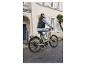 Femme conduisant un vélo électrique beige dans une rue pavée.
