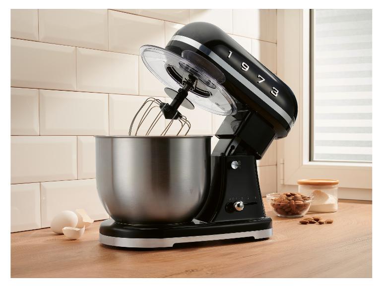 Robot pâtissier noir avec bol en acier inoxydable, œufs et amandes sur un comptoir en bois.