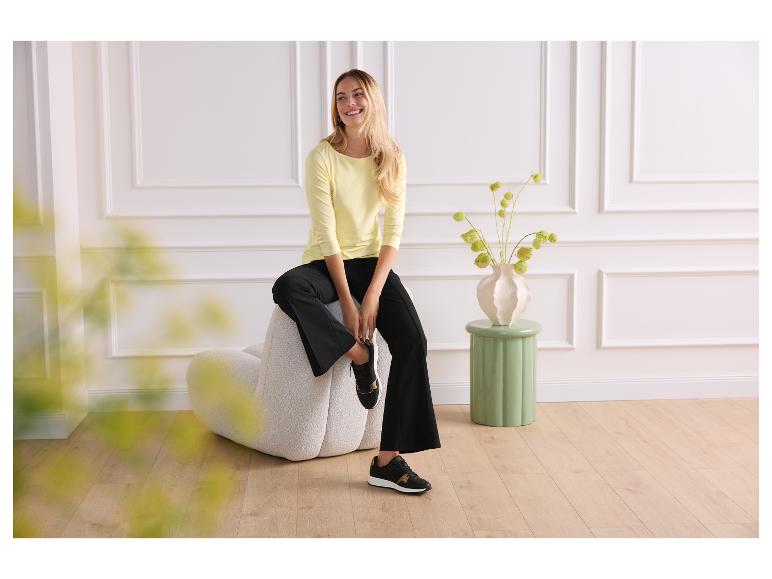 Femme en chemise jaune et pantalon noir assise sur une chaise beige.