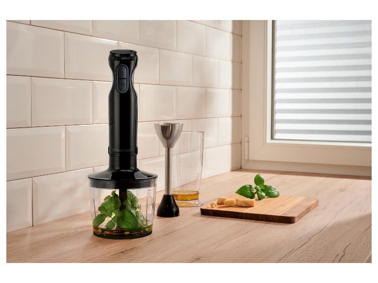 Mixeur plongeant avec verre doseur, basilic, fromage et huile d'olive sur plan de travail en bois.