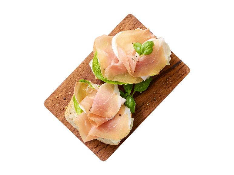 Deux tartines avec du prosciutto, de la mozzarella, de la laitue et du basilic sur une planche en bois.