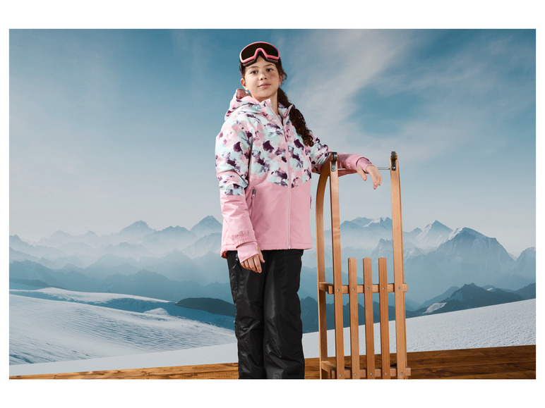 Une fille avec une veste de ski rose et un pantalon de ski noir se tient près d'une luge dans un paysage hivernal.