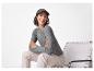 Femme souriante en pull gris et pantalon beige, assise sur une chaise.