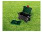 Chariot de jardin noir à roulettes avec couvercle vert, et plateau à outils vert à côté, sur l'herbe.