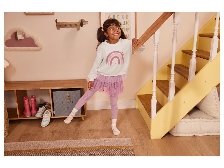 Fille joyeuse en sweat blanc arc-en-ciel et jupe en tulle rose près d'un escalier.