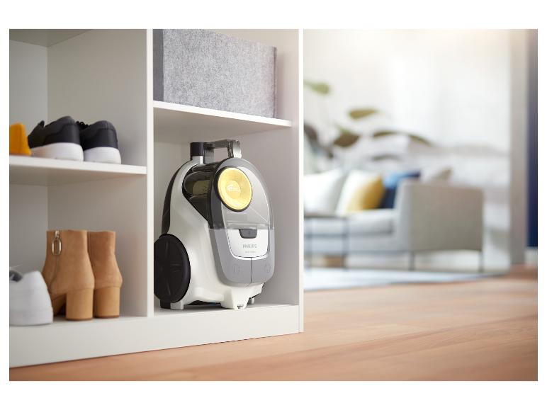 Aspirateur, chaussures et boîte de rangement sur une étagère blanche, avec salon flou en arrière-plan.