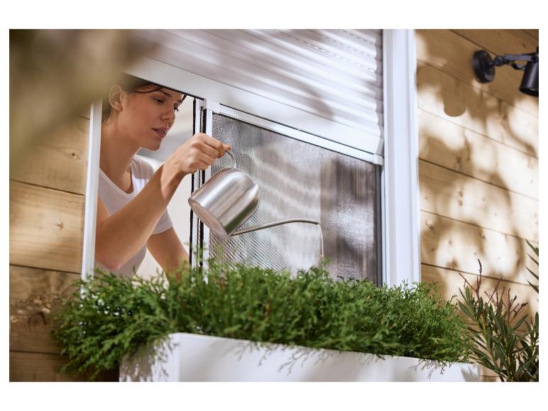 Femme arrosant des plantes dans une jardinière extérieure avec volet roulant et moustiquaire.