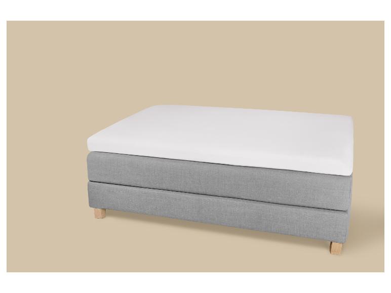 Sommier tapissier gris avec matelas et surmatelas blancs sur fond beige.