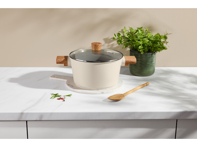 Casserole crème avec poignées en bois sur un plan de travail en marbre à côté d'un pot d'herbes.