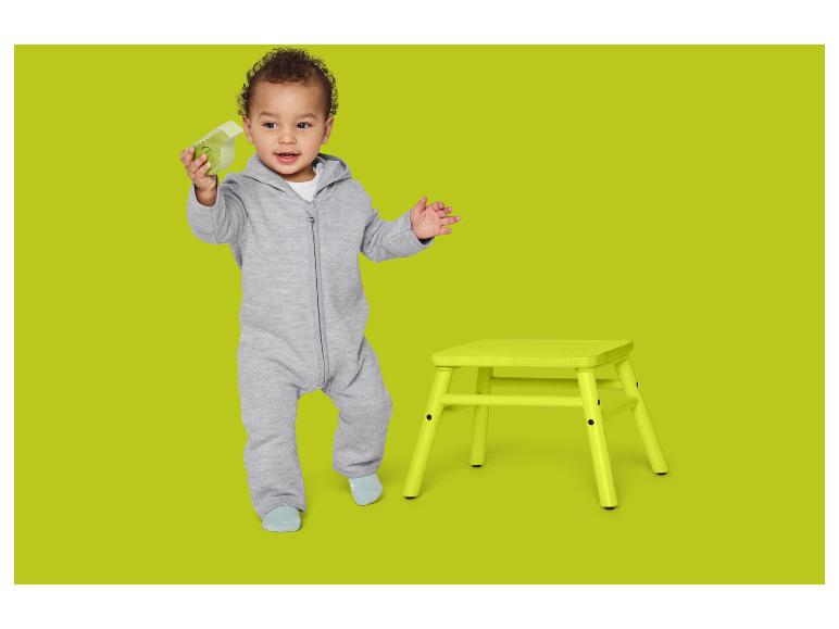 Bébé en grenouillère grise avec jouet vert et tabouret jaune sur fond vert.