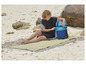 Une femme sur la plage avec un sac à dos isotherme Mistral et une serviette de plage rayée.