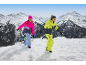 Deux enfants en combinaisons de ski et bonnets colorés, tirant une luge dans un paysage montagneux enneigé.