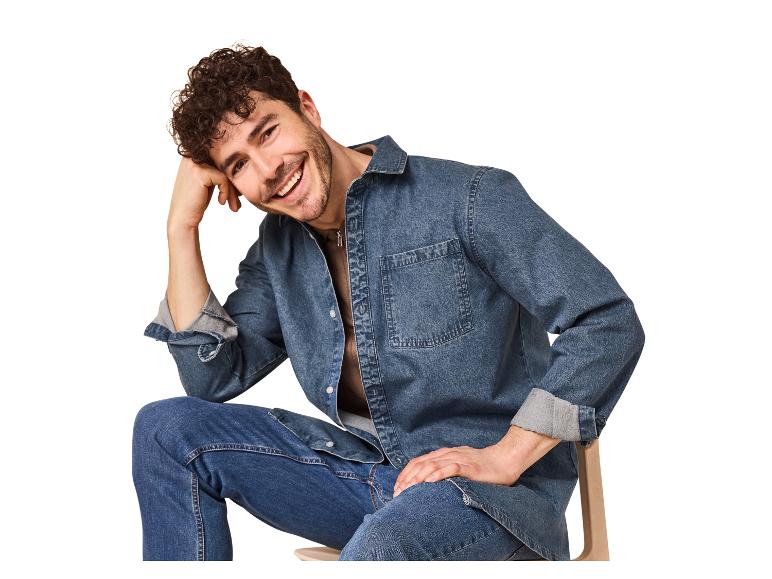 Homme souriant en chemise et jean en denim, assis sur une chaise.