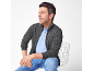 Homme en veste tricotée grise, polo bleu clair et jean, assis sur une chaise blanche.