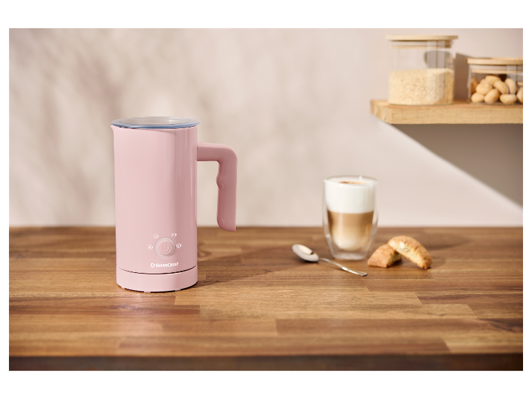 Un mousseur à lait rose SilverCrest sur une table en bois, à côté d'un cappuccino et de biscuits.