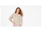 Femme aux cheveux bouclés portant un cardigan en maille ajourée beige et un pantalon beige.