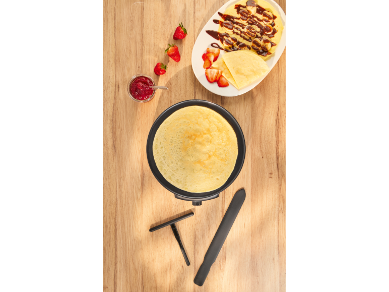 Crêpe dans une poêle, à côté d'une assiette de crêpes, bananes, fraises et confiture.