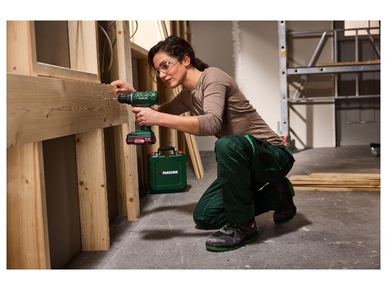 Femme en lunettes de sécurité et tenue de travail utilisant une perceuse sans fil sur un cadre en bois.