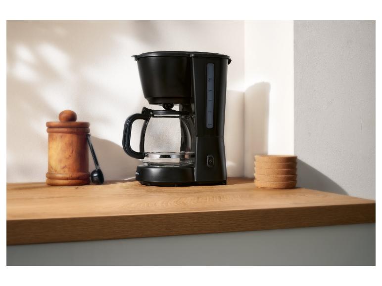 Cafetière filtre noire avec verseuse en verre, pot en bois et sous-verres en liège sur un comptoir.