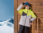 Un garçon porte une veste de ski et un bonnet avec des lunettes de ski, appuyé contre un mur en bois.