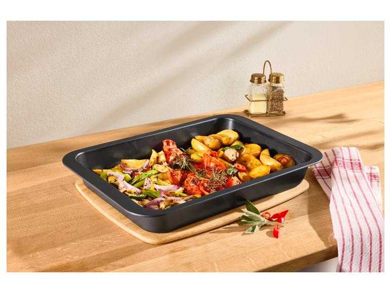 Plat de légumes rôtis avec pommes de terre, tomates et oignons dans un plat de cuisson noir sur une planche en bois.