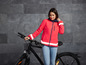 Une femme vêtue d'une veste rose vif avec des bandes réfléchissantes se tient à côté d'un vélo noir.