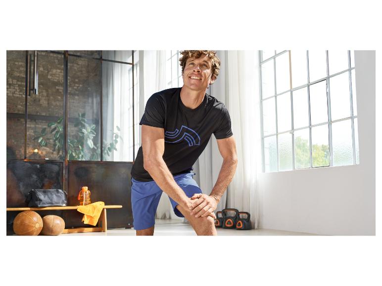 Homme en t-shirt de sport noir et short bleu s'étirant dans une salle de sport avec équipement.