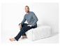 Un homme en pantalon de pyjama bleu et chemise grise assis sur un pouf blanc.