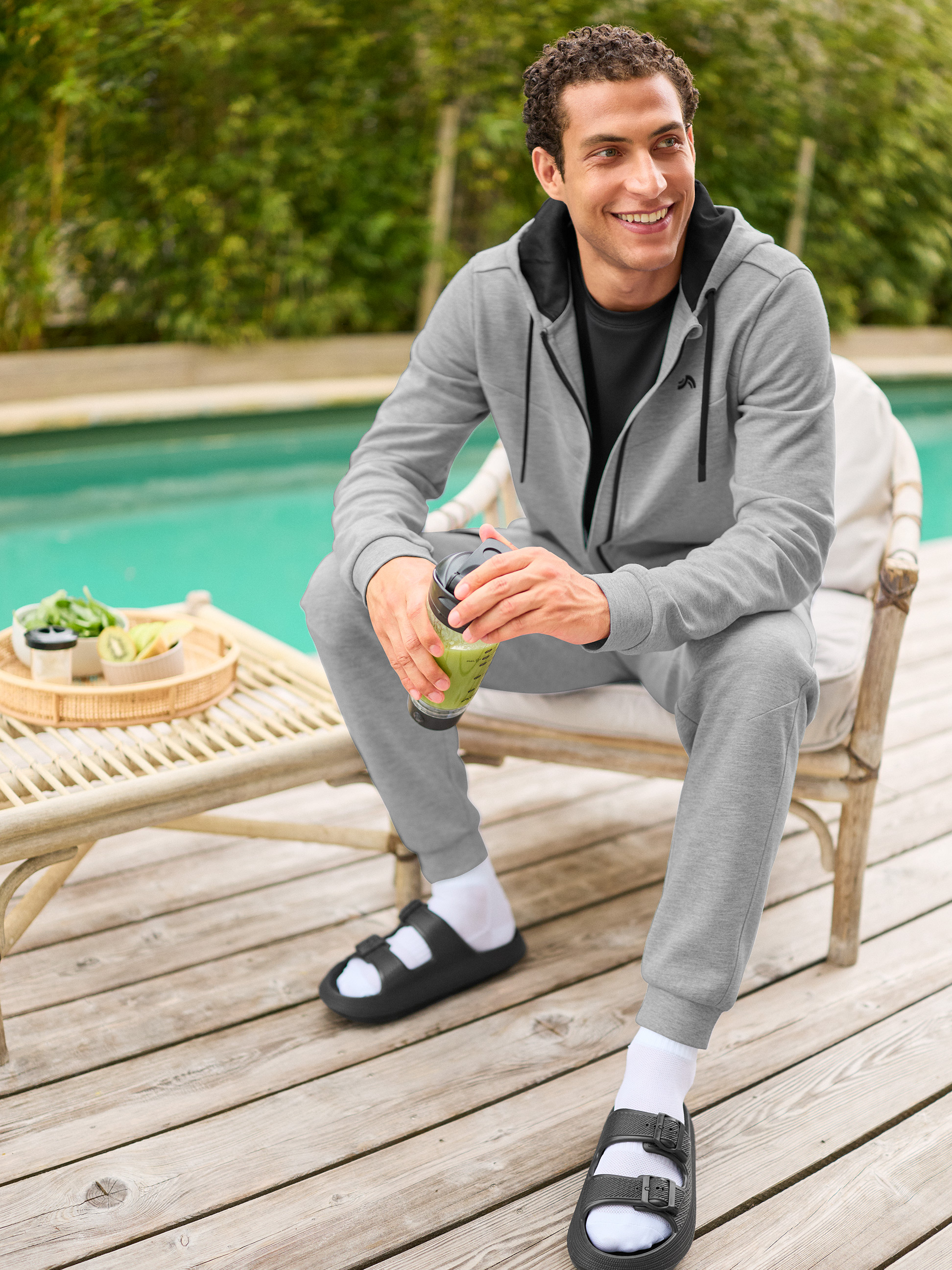 Homme en survêtement gris et sandales noires avec chaussettes blanches, tenant un smoothie vert au bord de la piscine.