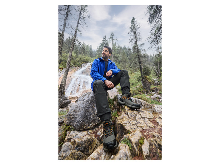 Un homme en veste bleue et en chaussures de randonnée marron assis sur des rochers dans une forêt.
