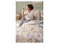 Femme en pyjama assise sur un lit avec une parure de lit florale, à côté d'une table de chevet avec des livres et une tasse.