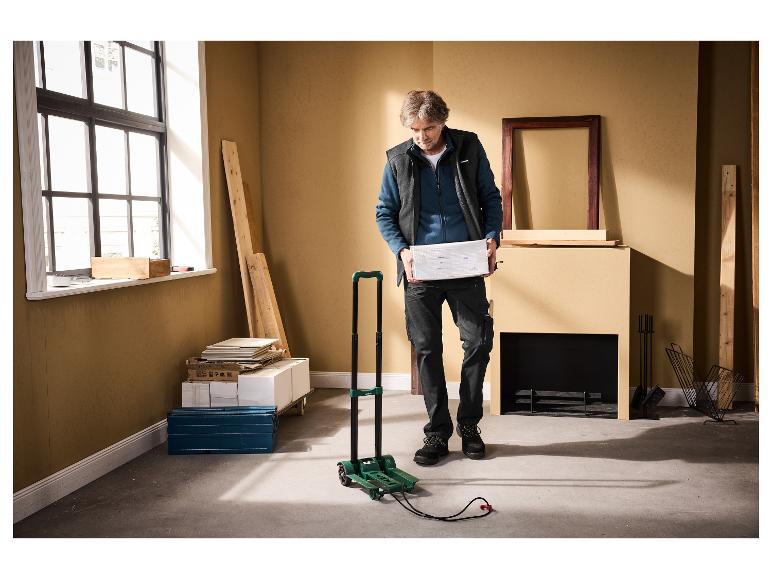 Homme tenant un colis, avec un chariot pliable et des matériaux de construction à proximité.