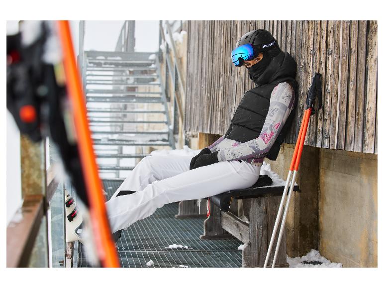 Une femme en tenue de ski, gilet noir et pantalon blanc, assise sur un banc.