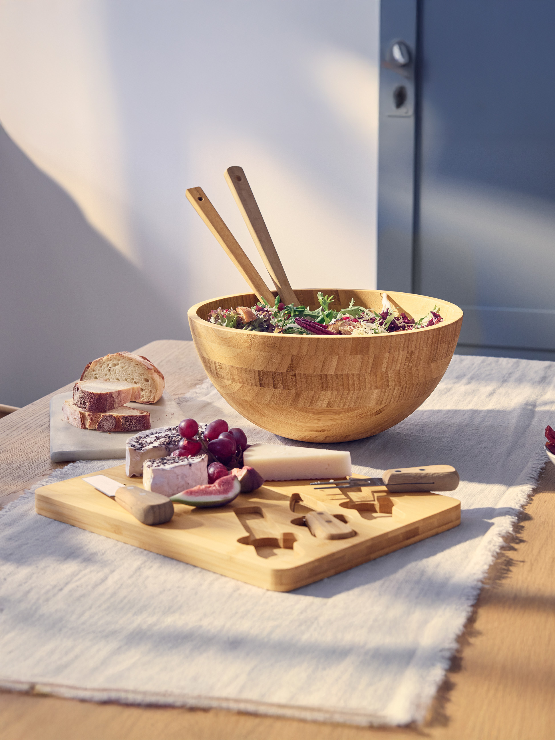 Salade dans un bol en bois avec ustensiles, plateau de fromages avec pain et fruits.