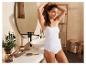 Une femme en débardeur blanc et en sous-vêtements, avec une brosse à cheveux sur le lavabo de la salle de bain.